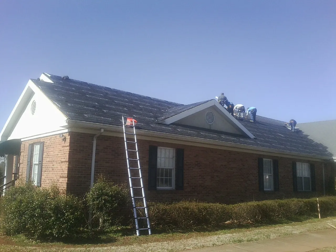 Crew tearing shingles off a brick ranch roof with a ladder set — Emergency Roof Repair work by our crew in Fort Cobb