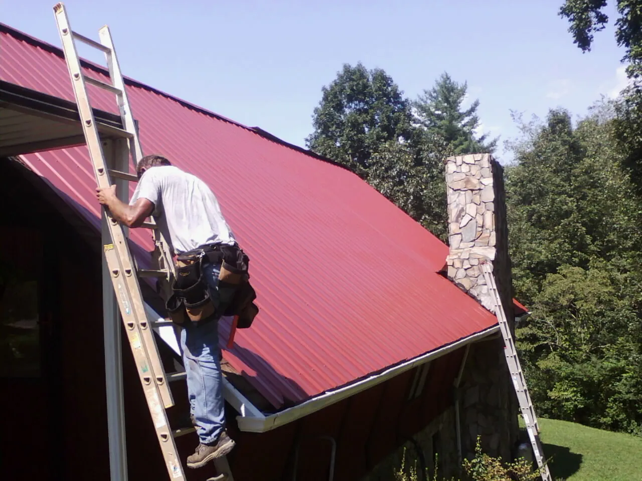 Roofer on a ladder beside a red metal roof and stone chimney — Soffit & Fascia Repair by Roofing Brothers in Fort Cobb